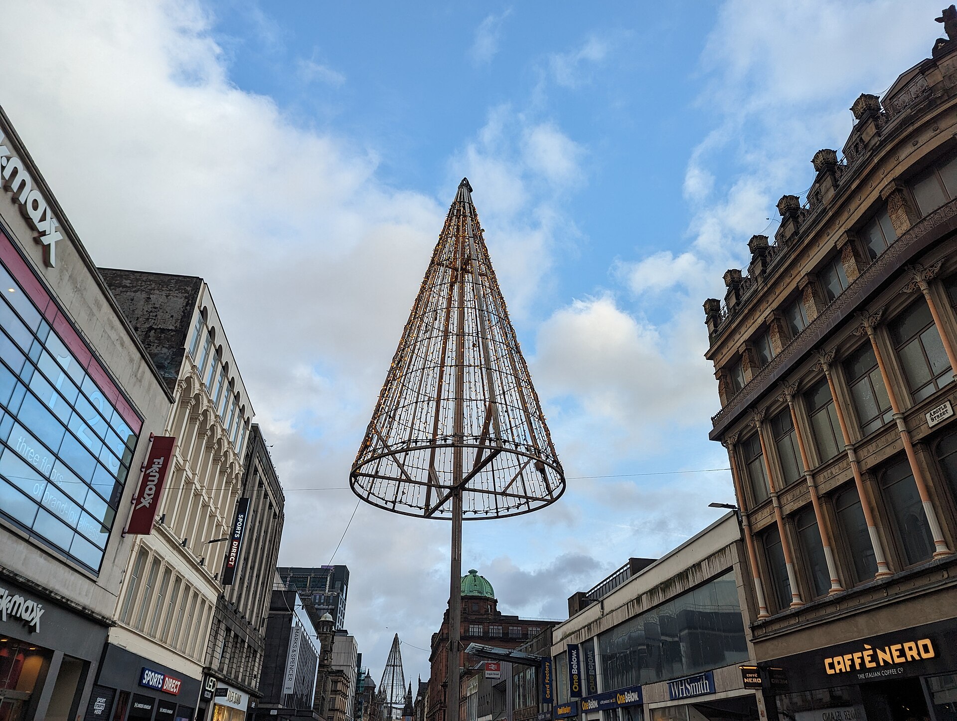 A tall cone-shaped Christmas light tree on a pole above Argyle Street, with shops on both sides and a second light tree in the distance under a blue sky