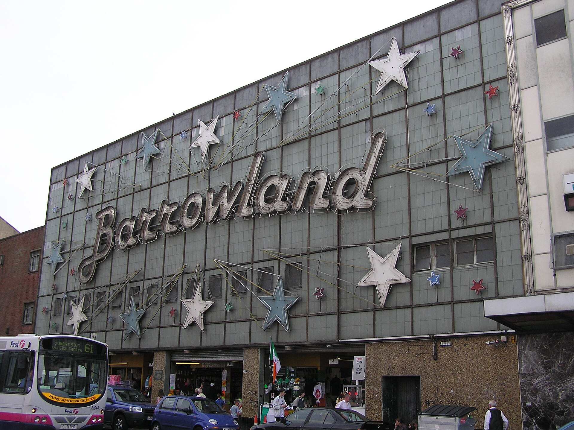 The Barrowland building by day, with the unlit script sign and stars over open market stalls on the Gallowgate and a bus passing in front