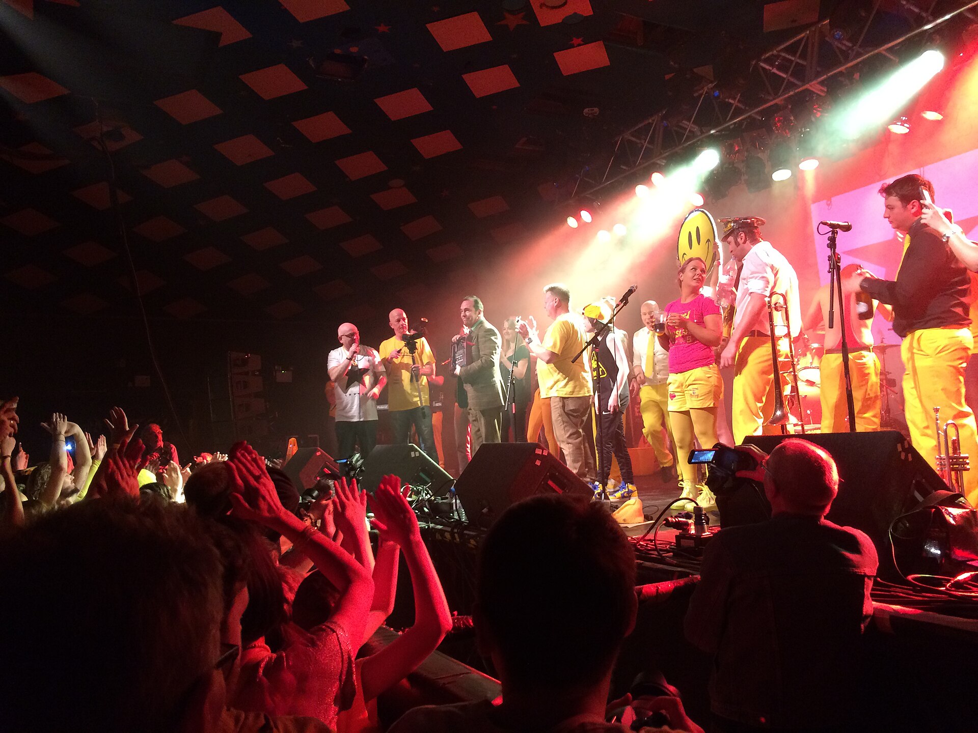 Performers in yellow costumes crowd the Barrowland stage during a Hall of Fame presentation, with fans' raised hands in the foreground and the checkered star-dotted ceiling above