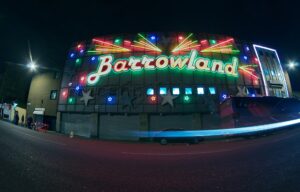 The Barrowland neon script sign and coloured stars lit up at night on the Gallowgate, with a car's light trail passing below