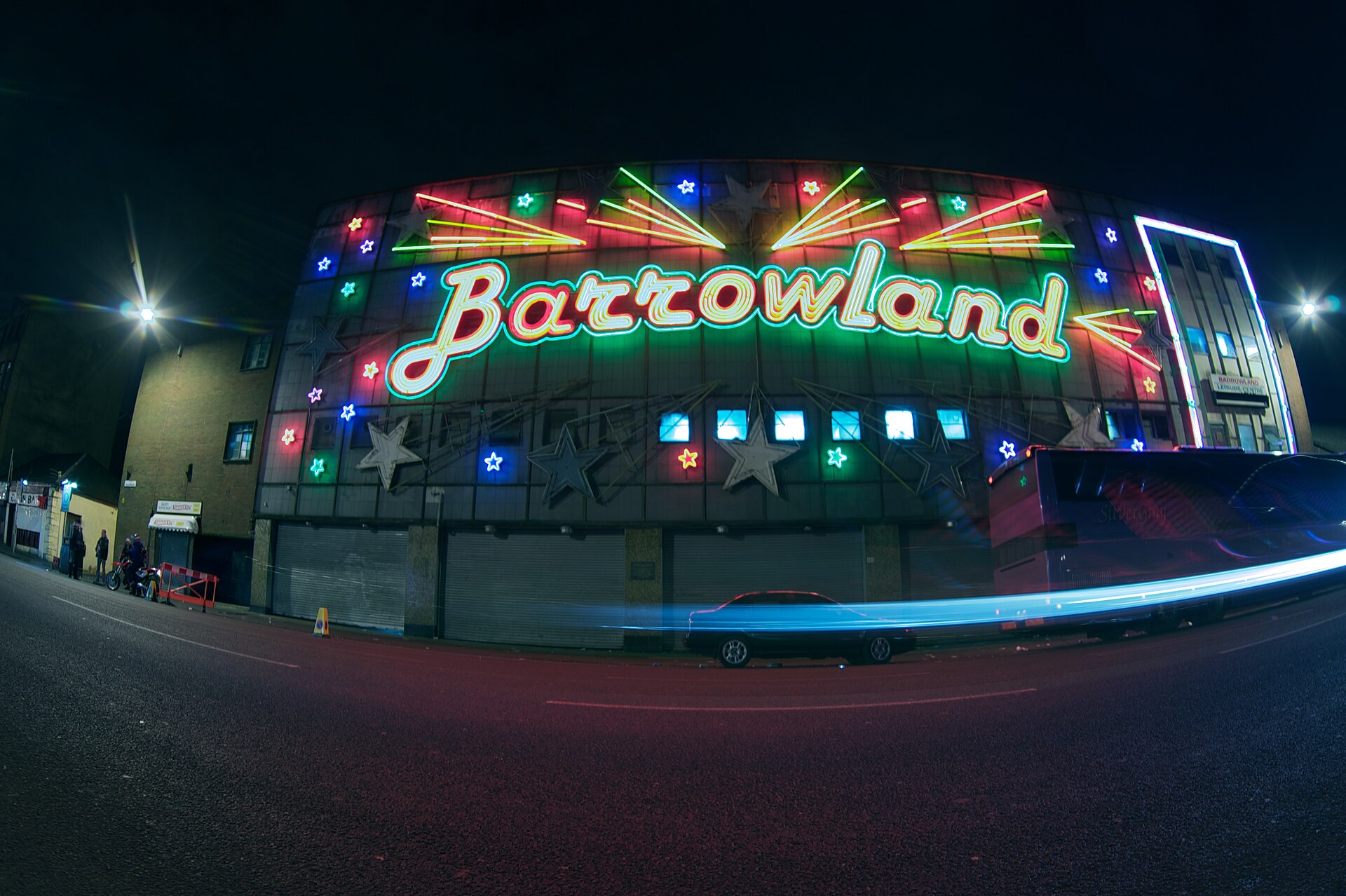 The Barrowland neon script sign and coloured stars lit up at night on the Gallowgate, with a car's light trail passing below