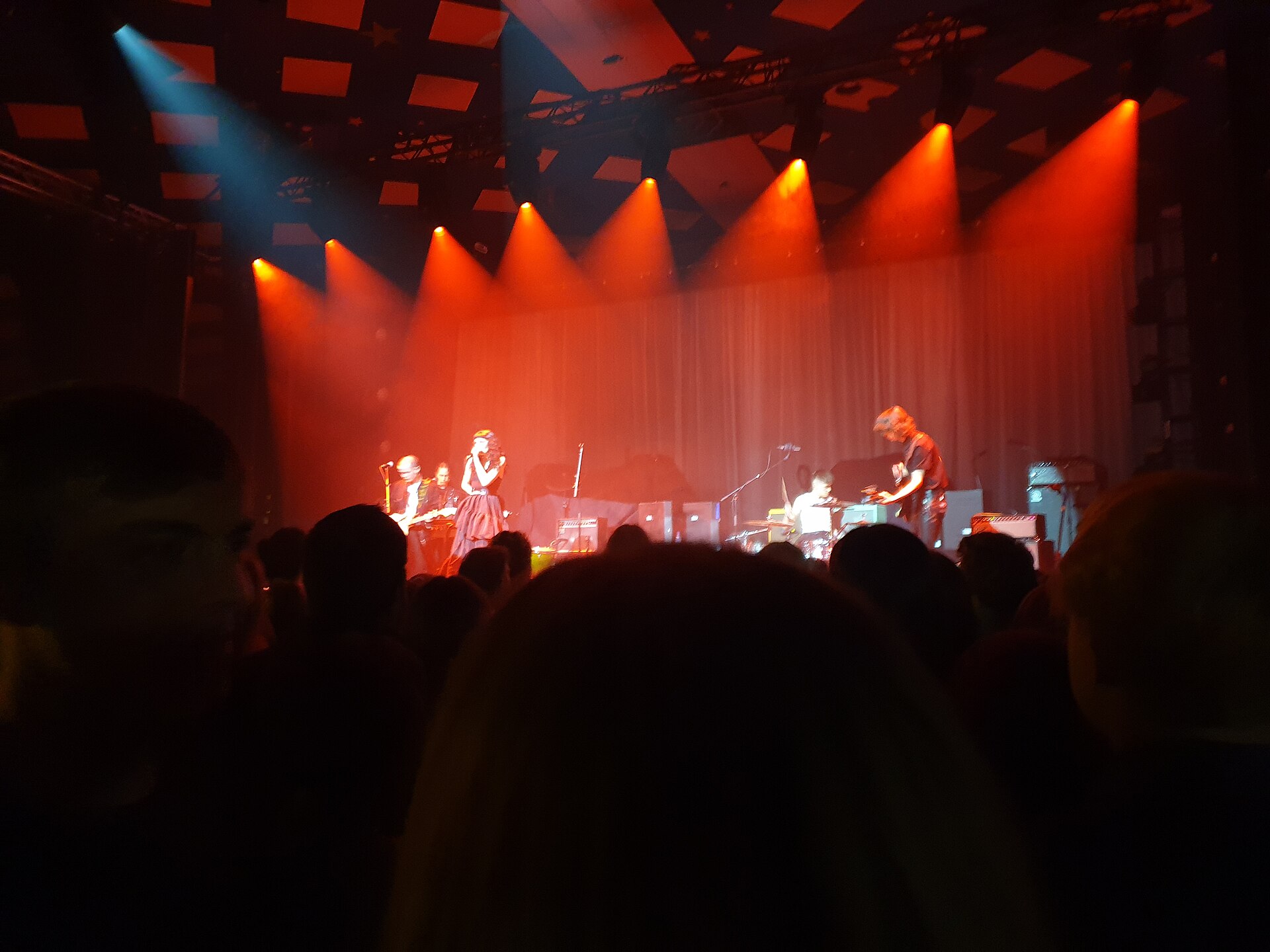 A band on the Barrowland stage lit in red and orange, seen over the heads of a standing crowd, with the square ceiling tiles and small stars overhead
