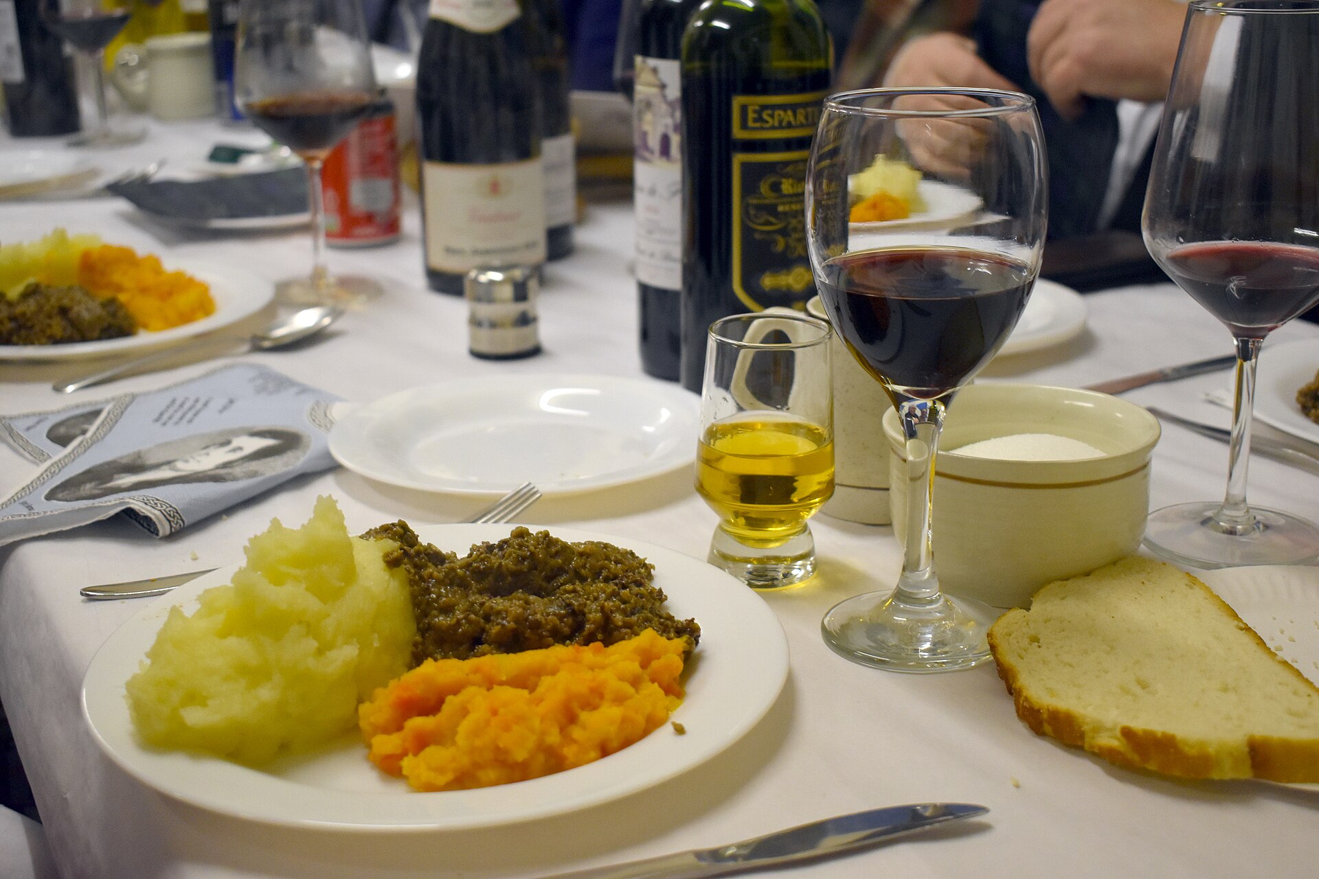 A plate of haggis, mashed potato and mashed swede on a white tablecloth, with a glass of whisky, red wine, bread and a napkin printed with a portrait of Robert Burns