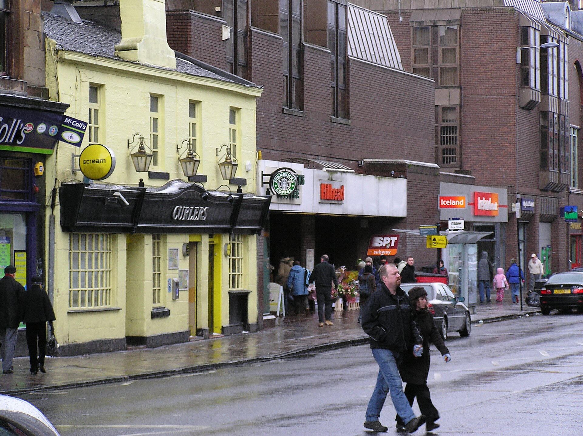 A wet Byres Road with shoppers crossing beside a pale yellow corner pub and a row of shops