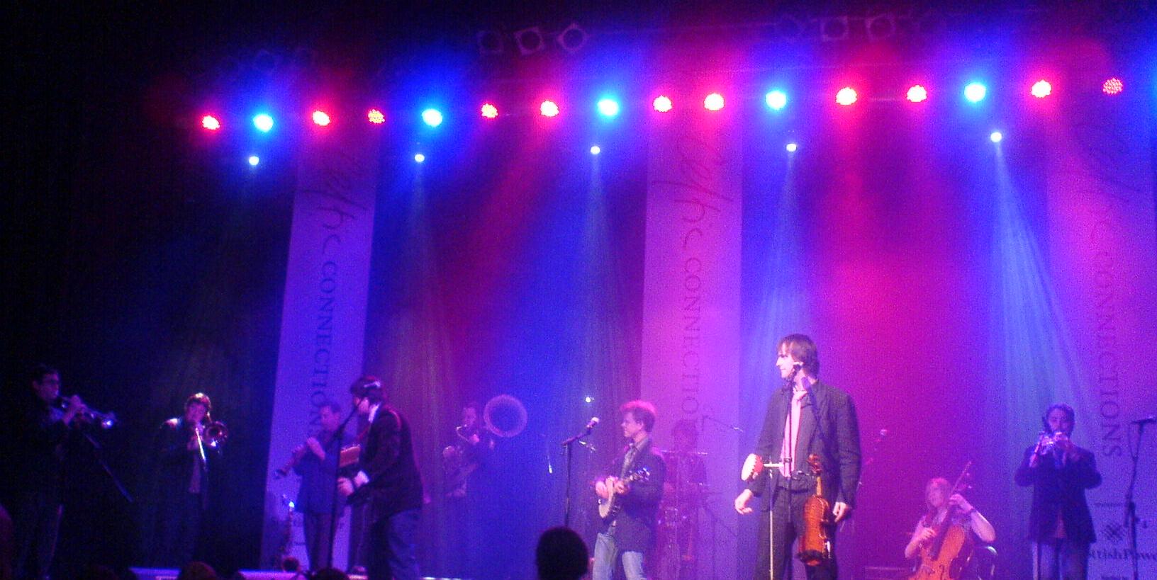 A large folk band with fiddles, brass, banjo and cello on a stage lit in pink and blue, with Celtic Connections banners hanging behind
