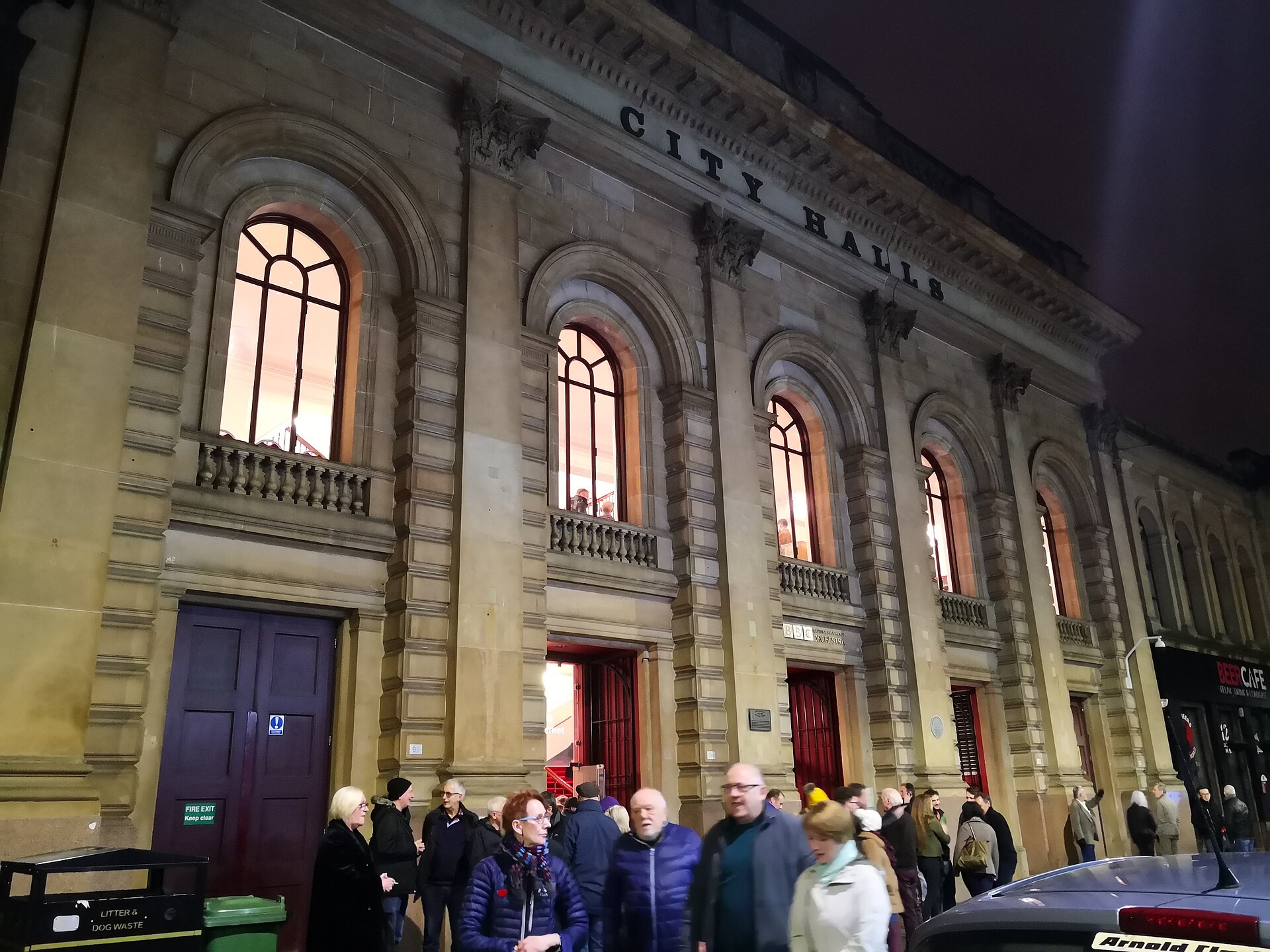 The floodlit sandstone front of City Halls on Candleriggs at night, with tall arched windows lit from inside and a crowd gathering at the door