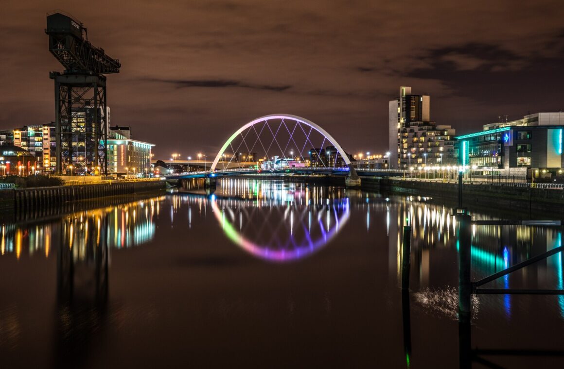 The Clyde Arc bridge lit in rainbow colours and the Finnieston Crane at night, with city lights reflected in the still River Clyde
