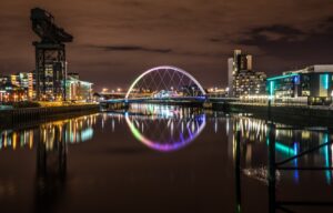 The Clyde Arc bridge lit in rainbow colours and the Finnieston Crane at night, with city lights reflected in the still River Clyde
