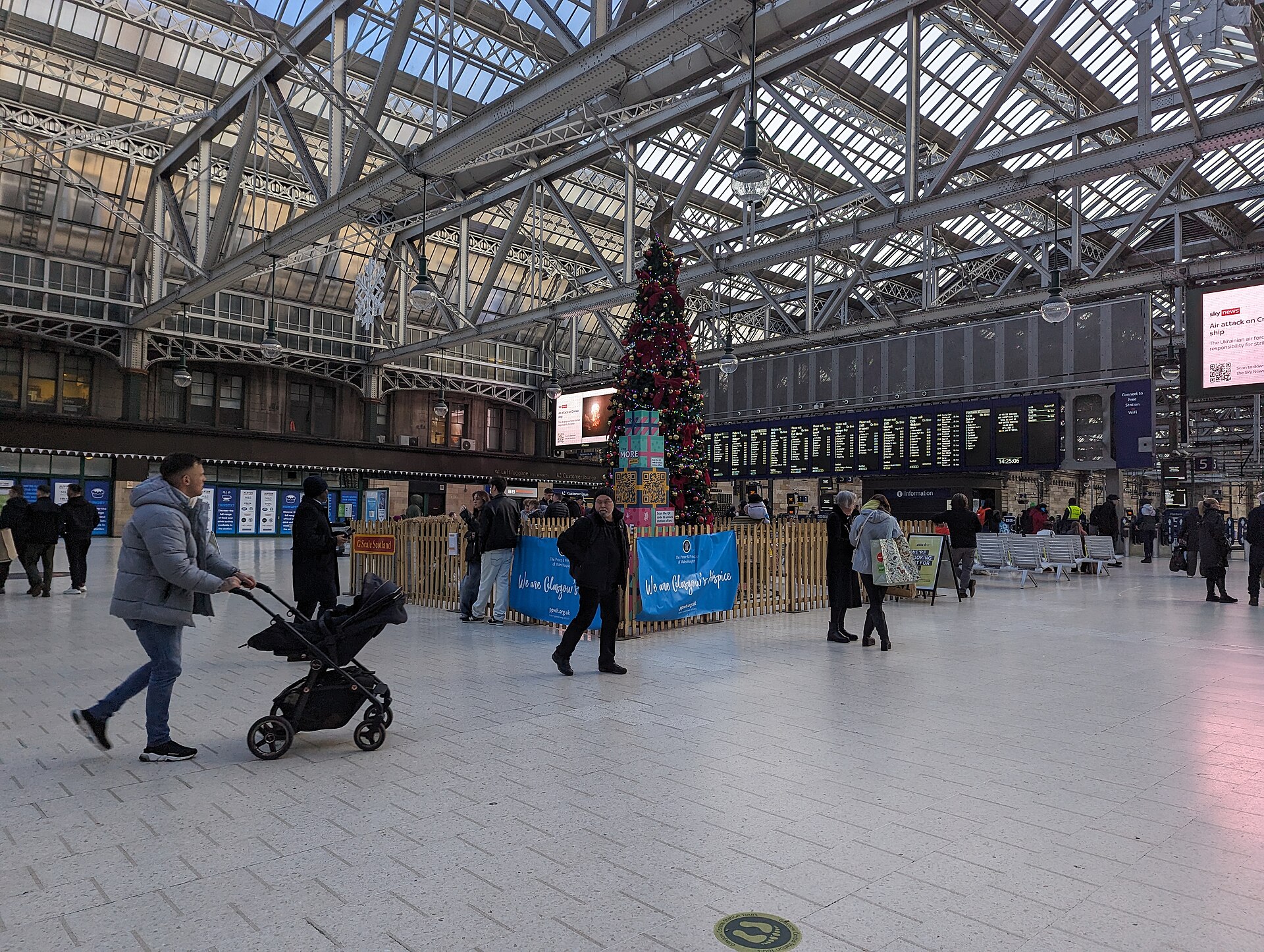 The concourse of Glasgow Central station under its iron and glass roof, with a decorated Christmas tree in front of the departure boards and people crossing the floor