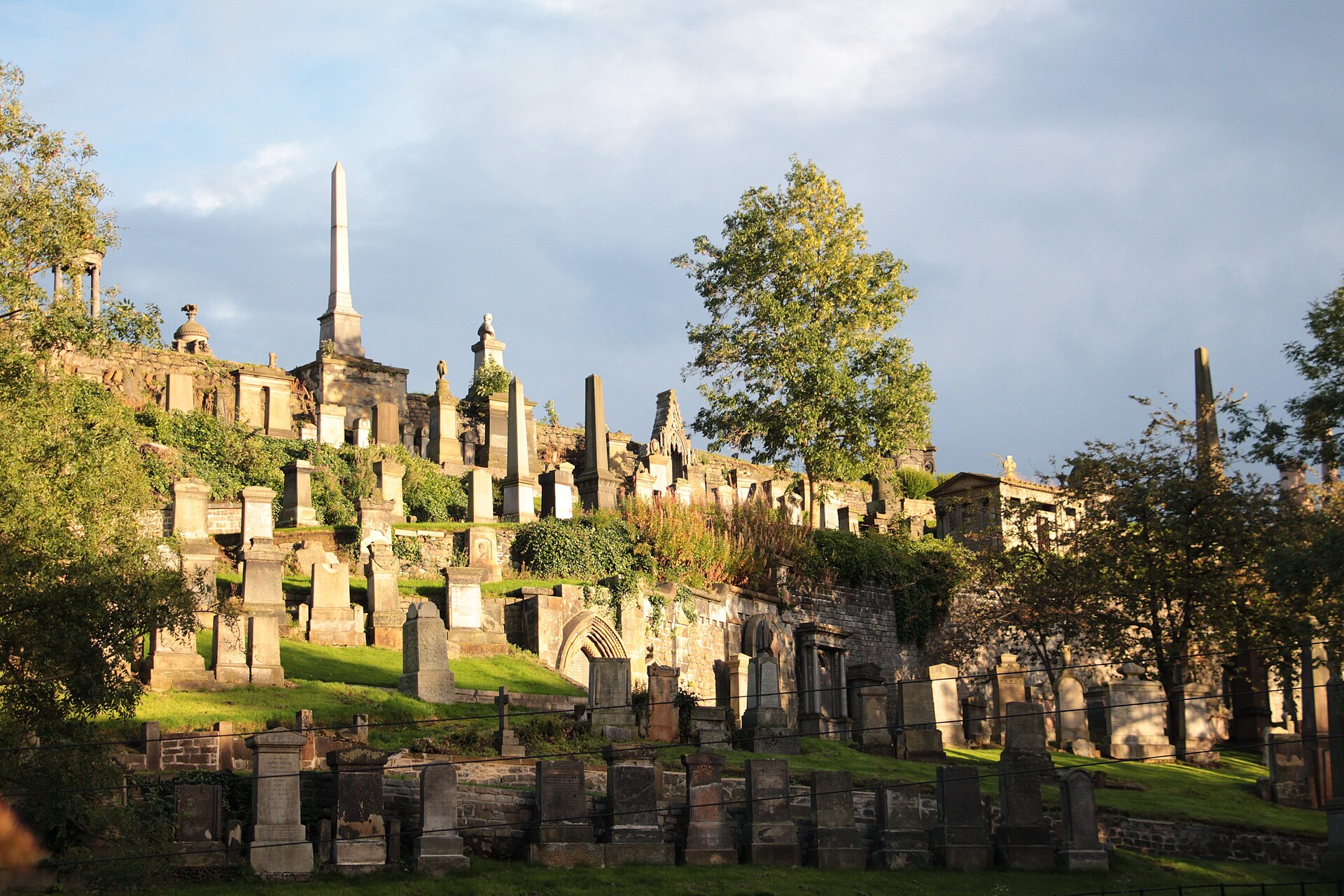 Victorian monuments, obelisks and a domed memorial climbing the terraced hillside of the Glasgow Necropolis in low evening sun