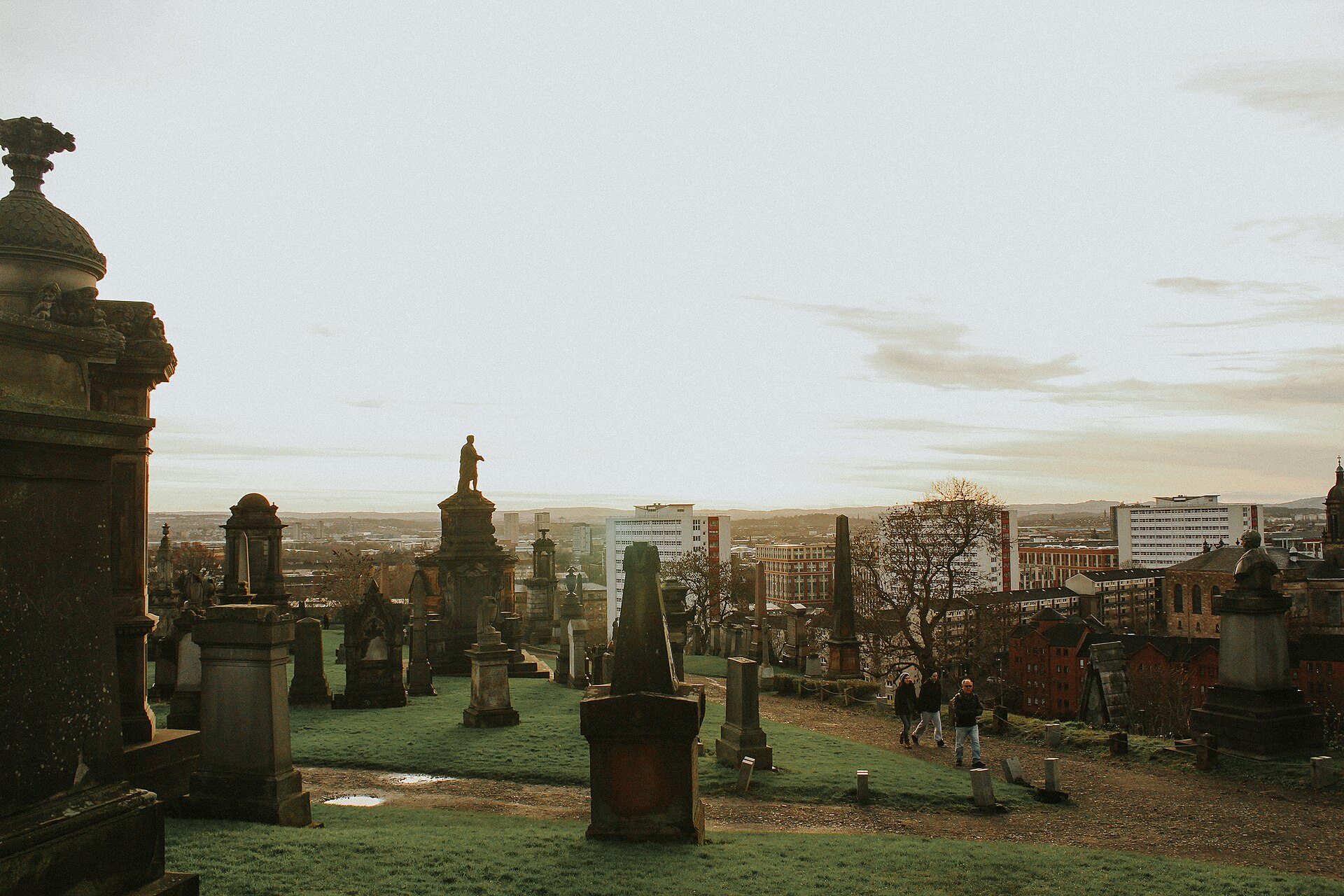 Walkers on a gravel path between Victorian monuments and a statue on top of the Glasgow Necropolis, with tower blocks and hills on the horizon under a pale winter sky