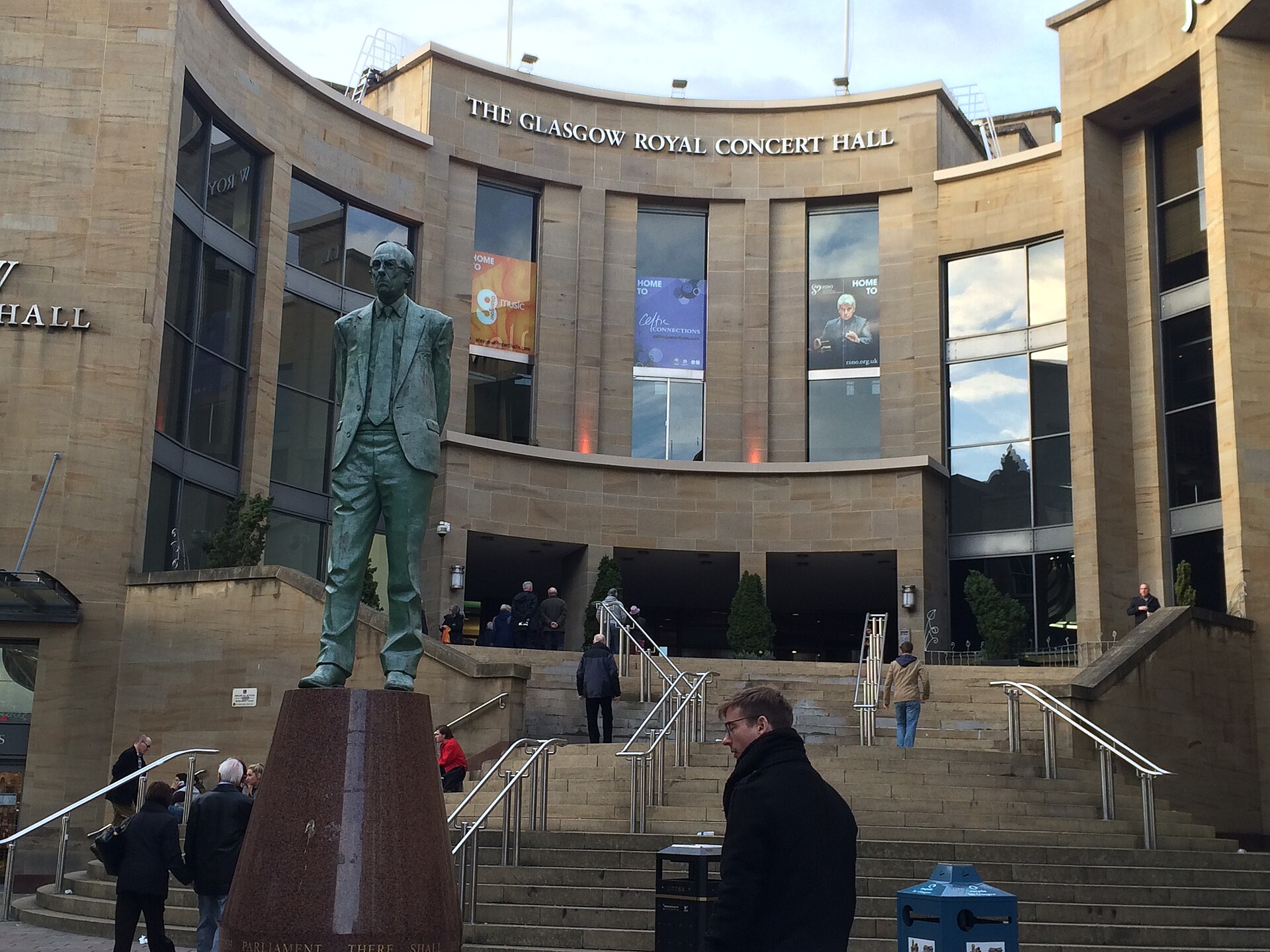 The curved sandstone front of the Glasgow Royal Concert Hall behind the bronze statue of Donald Dewar, with people walking up the wide steps