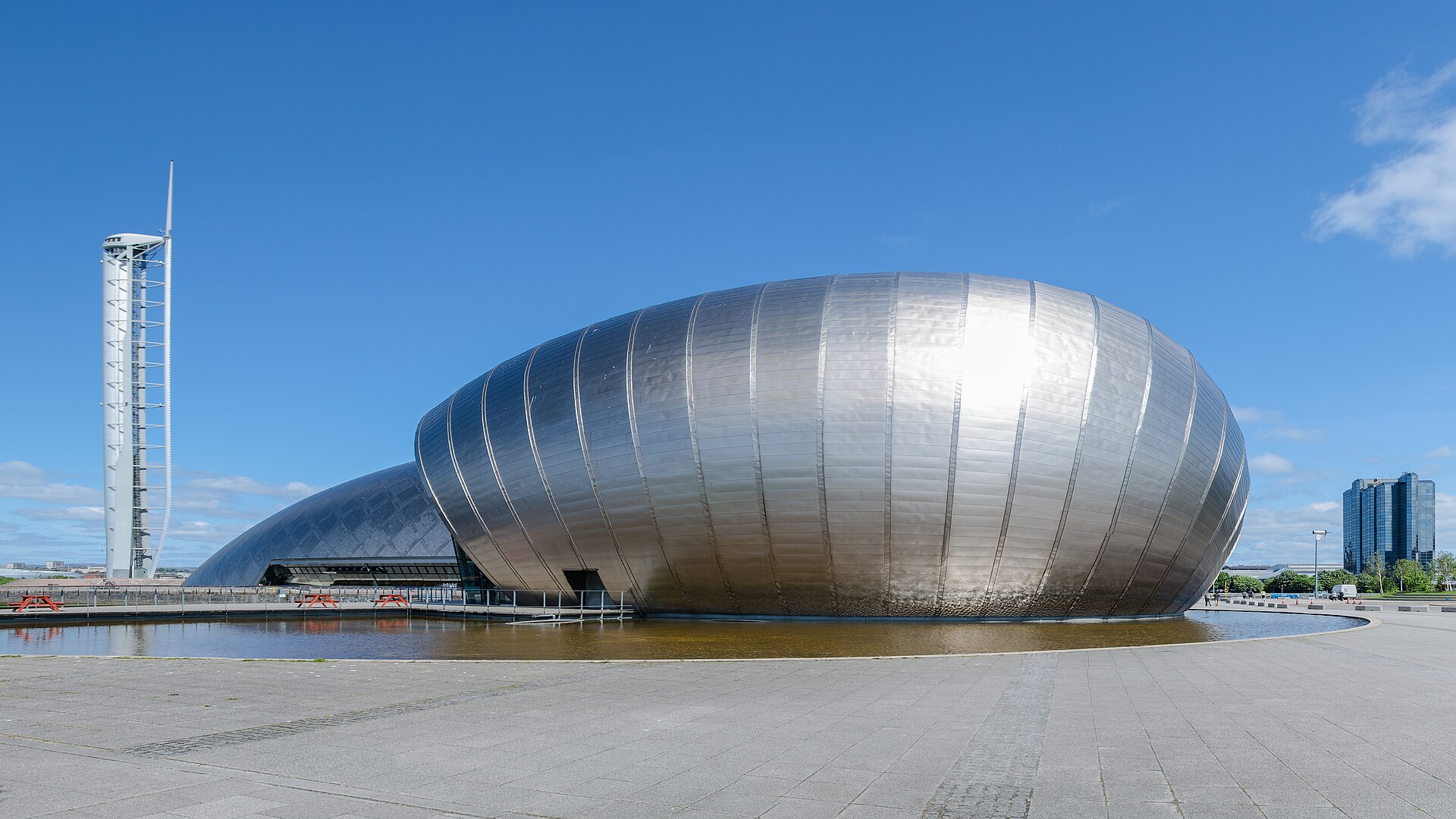 The curved titanium-clad buildings of the Glasgow Science Centre beside a pool on Pacific Quay, with the white Glasgow Tower rising on the left under a blue sky