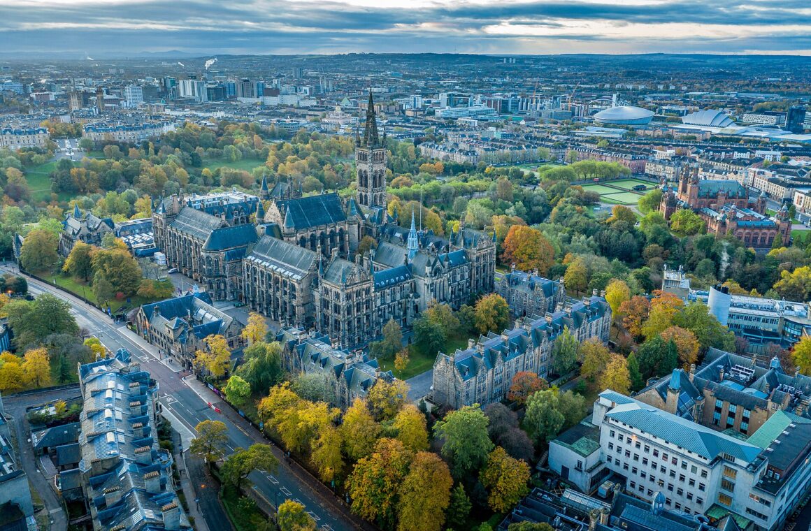 Aerial view of the University of Glasgow's Gothic main building among autumn trees, with Kelvingrove Art Gallery to the right and the city centre, SEC Armadillo and OVO Hydro beyond
