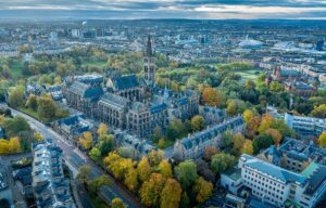 Aerial view of the University of Glasgow's Gothic main building among autumn trees, with Kelvingrove Art Gallery to the right and the city centre, SEC Armadillo and OVO Hydro beyond