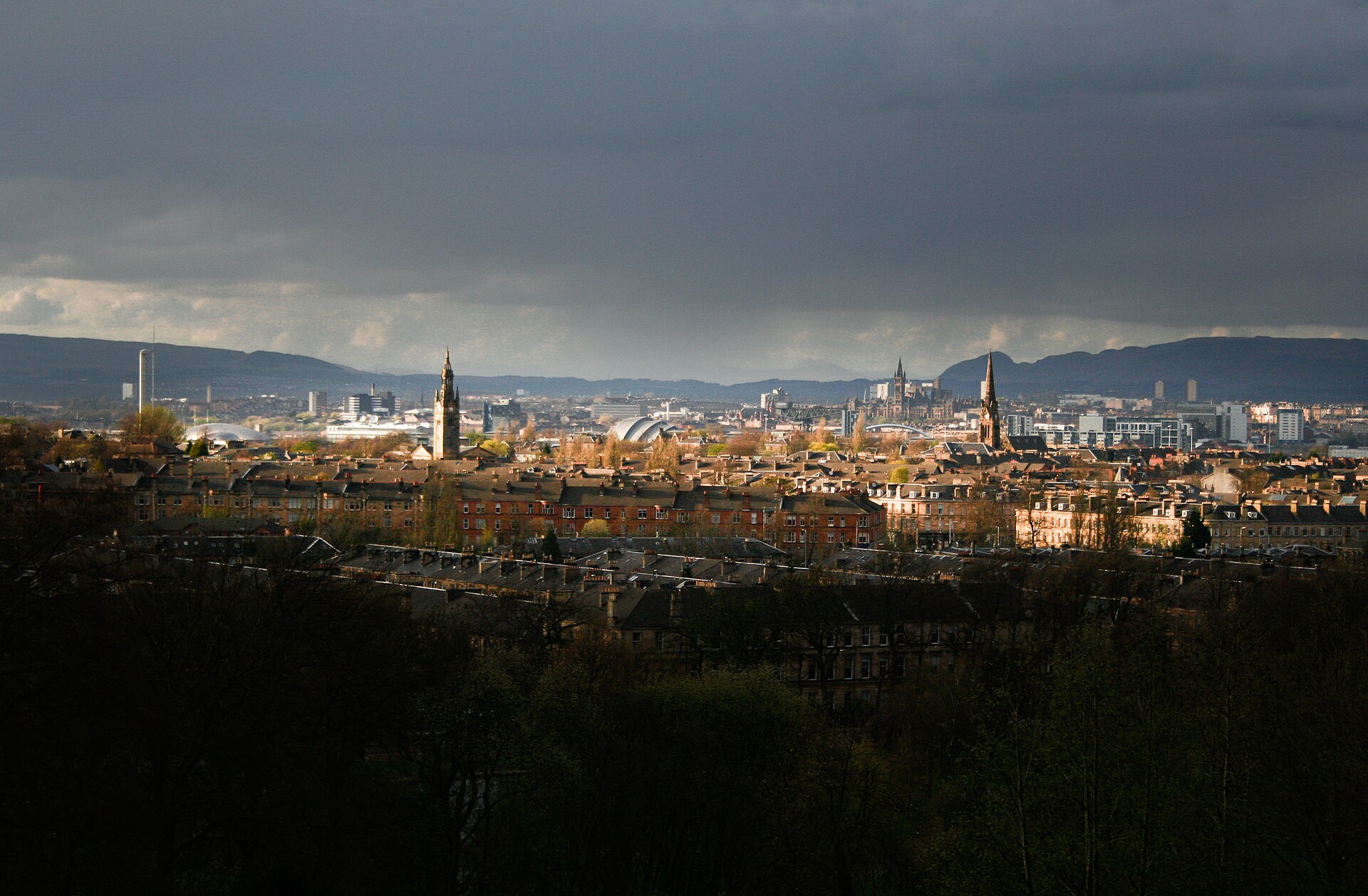 Sunlight breaking under dark storm clouds across Glasgow's sandstone rooftops and spires, with the SEC Armadillo, the University of Glasgow tower and the Campsie Fells in the distance