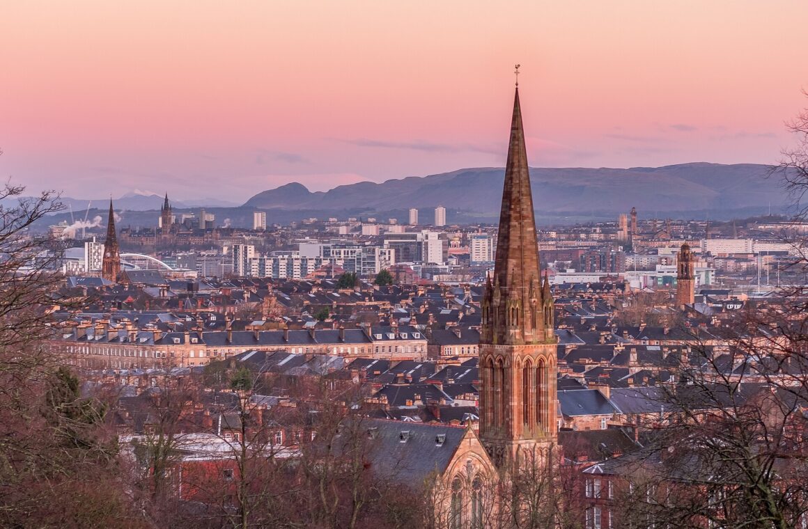 A pink winter dawn over Glasgow's sandstone tenement rooftops and church spires, with bare trees in front and the University of Glasgow tower and the Campsie Fells in the distance