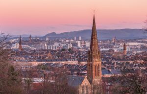A pink winter dawn over Glasgow's sandstone tenement rooftops and church spires, with bare trees in front and the University of Glasgow tower and the Campsie Fells in the distance