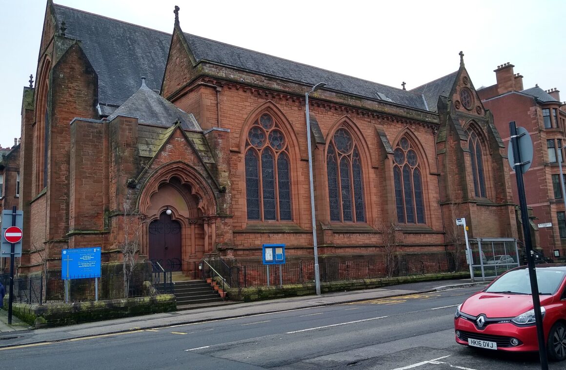 The red sandstone Gothic Hyndland Parish Church on a street corner, with arched windows and a red car parked outside
