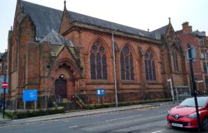 The red sandstone Gothic Hyndland Parish Church on a street corner, with arched windows and a red car parked outside