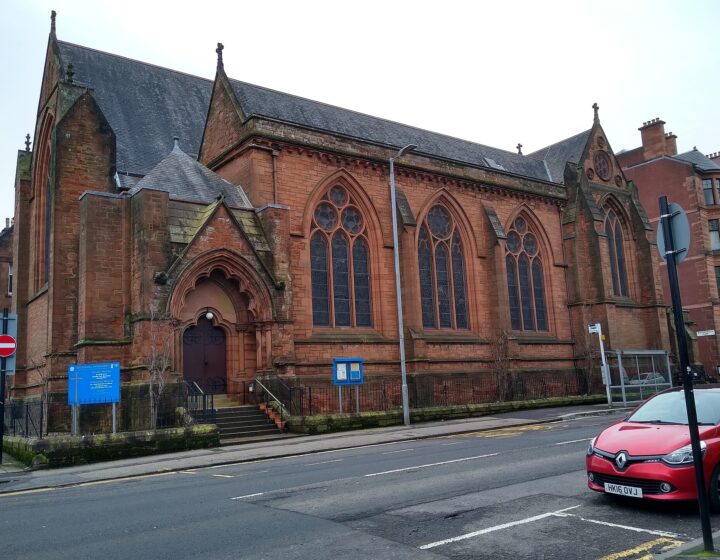 The red sandstone Gothic Hyndland Parish Church on a street corner, with arched windows and a red car parked outside