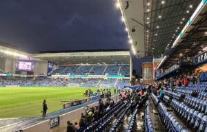 Ibrox Stadium filling up before an evening kick-off, seen from a low seat with the Copland Stand opposite
