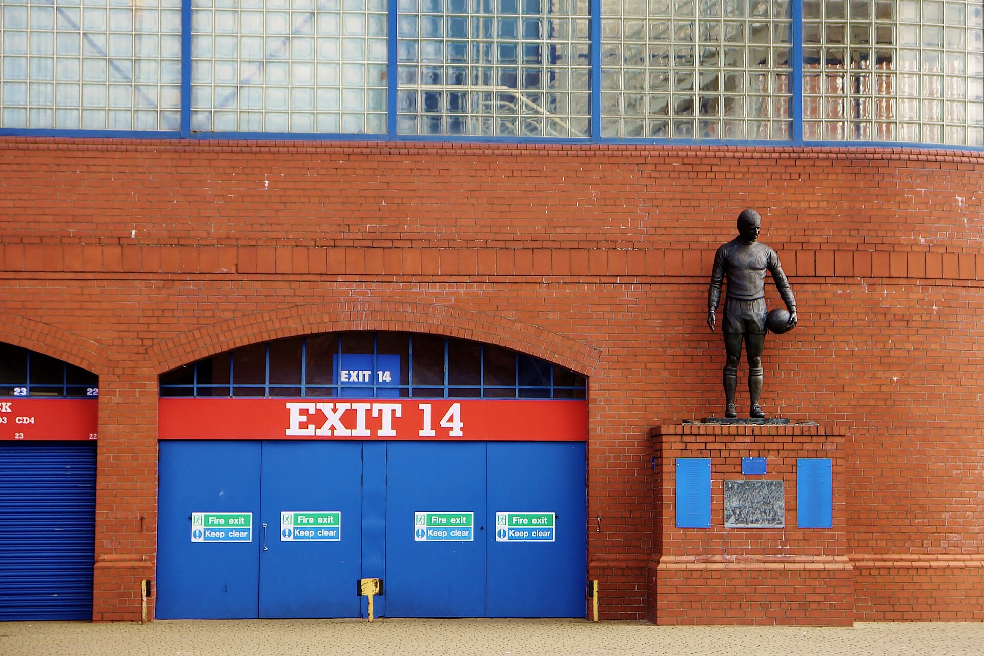 The bronze John Greig statue standing on a brick plinth beside Exit 14 on the red-brick wall of the Ibrox Main Stand