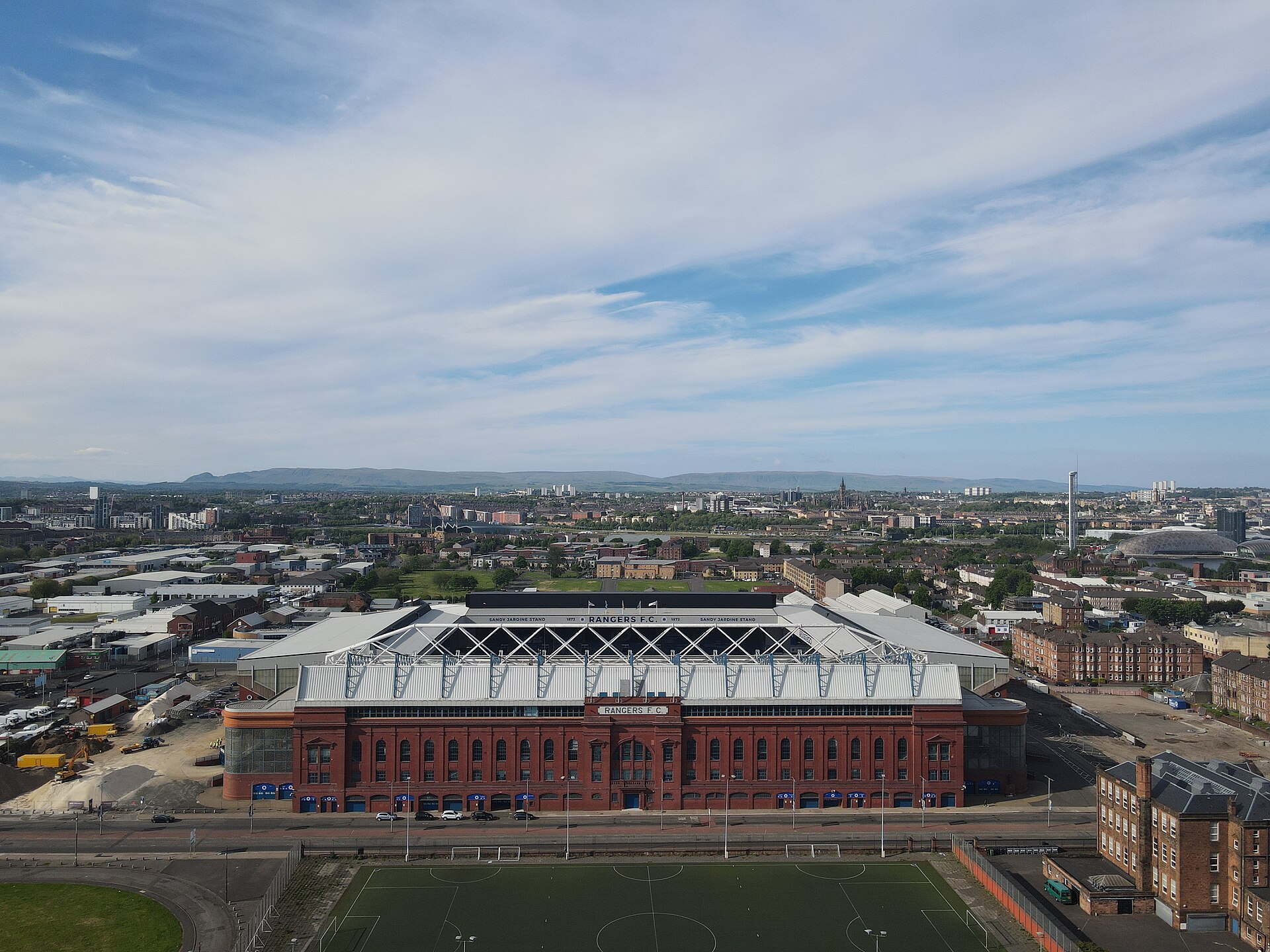 Aerial view of Ibrox Stadium in Govan, showing the red-brick Bill Struth Main Stand facade with the Glasgow skyline behind