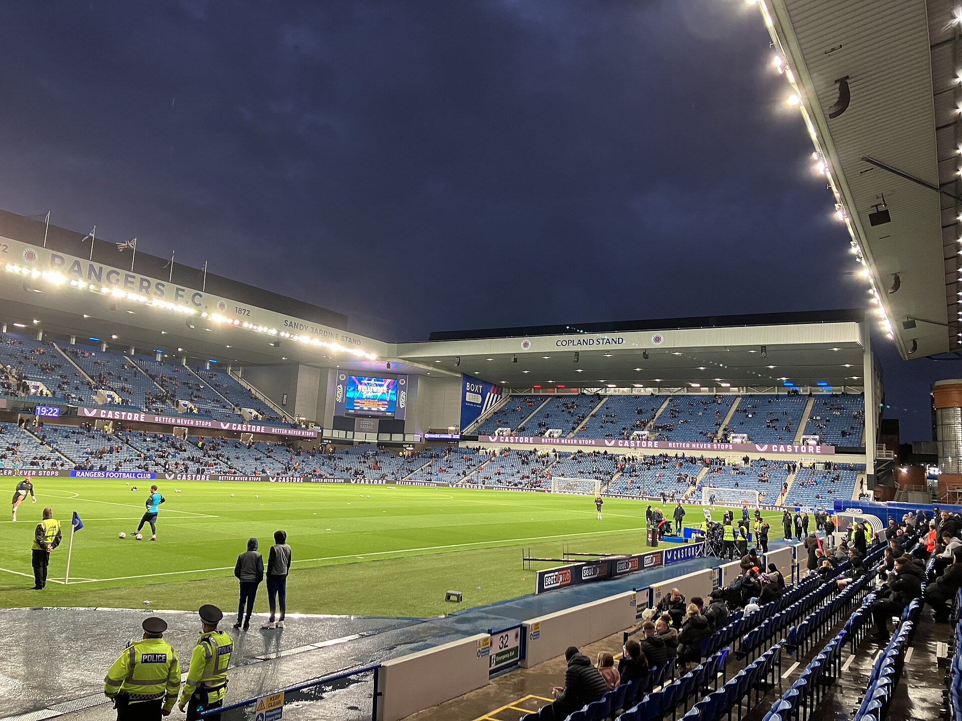 Ibrox Stadium under floodlights before an evening kick-off, with the Sandy Jardine Stand and Copland Stand lit and players warming up