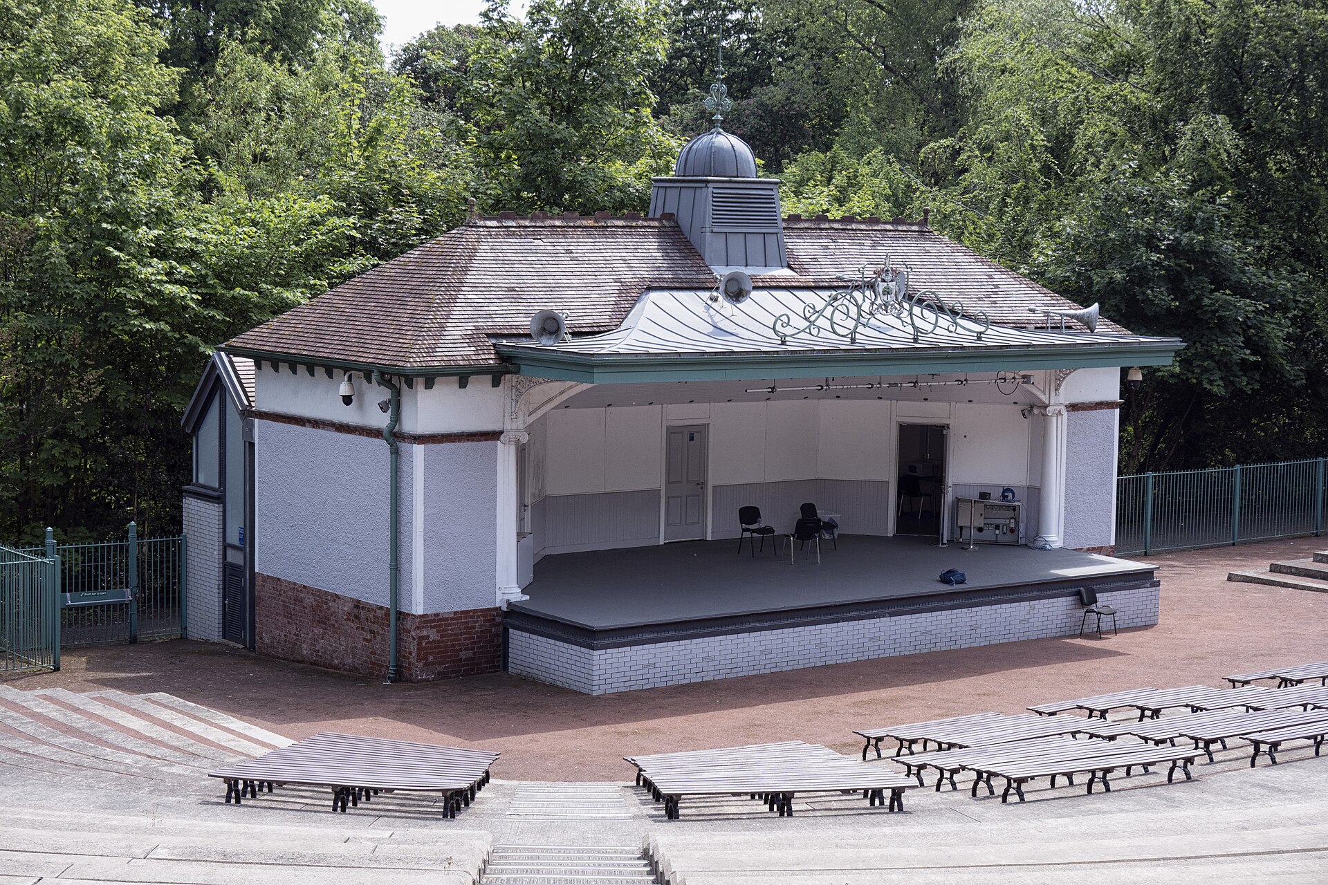 The restored bandstand in Kelvingrove Park, with an ornate roof, surrounded by trees in full summer leaf and rows of wooden benches in the amphitheatre