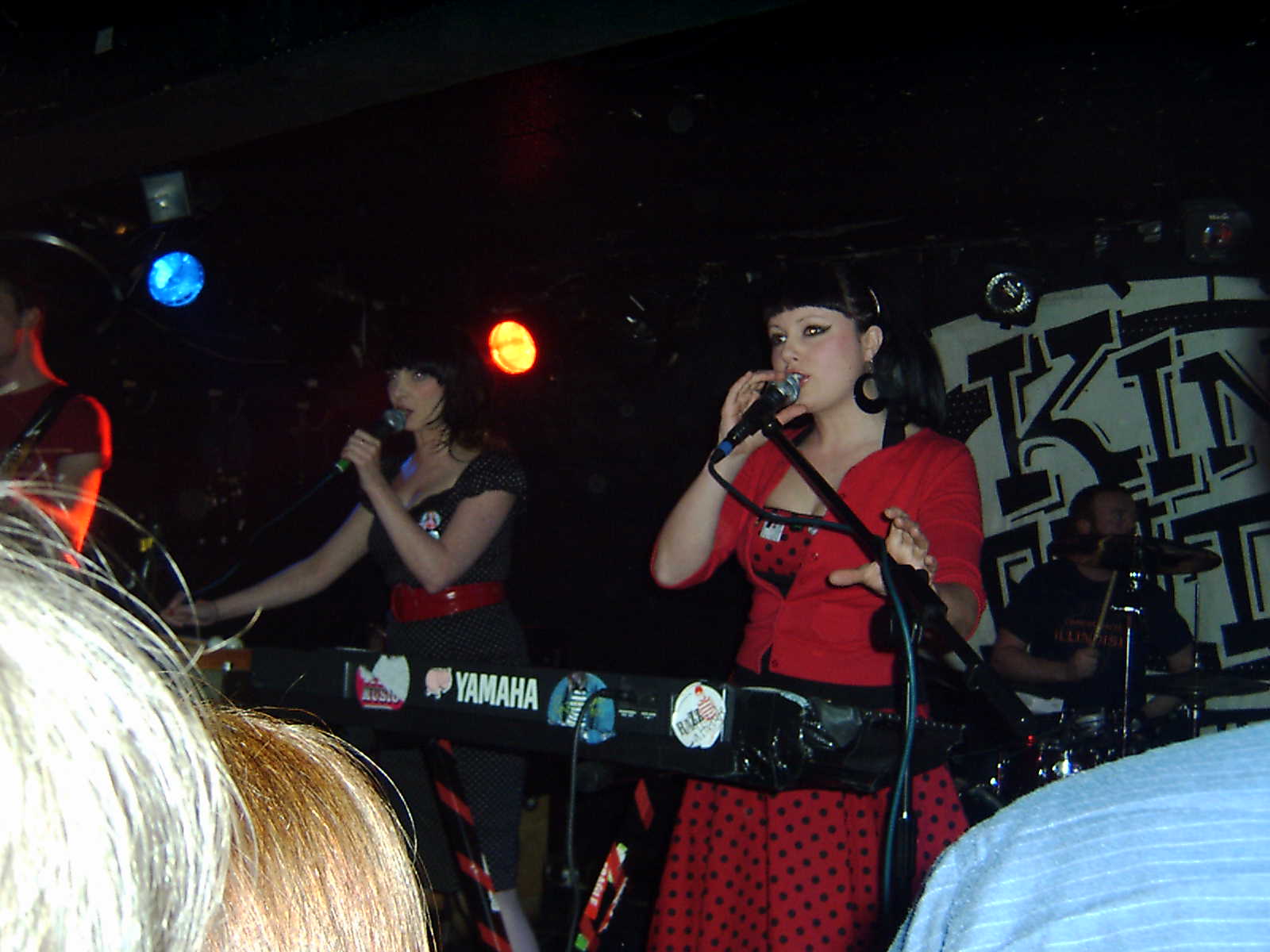 Two singers in red and black polka-dot outfits behind a stickered keyboard at King Tut's, with the venue's black and white lettering on the wall behind
