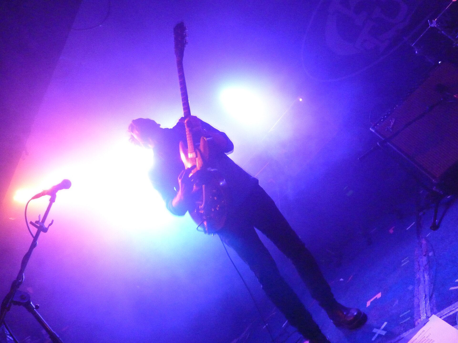 A guitarist silhouetted in blue and pink stage light, photographed from below at the edge of the King Tut's stage