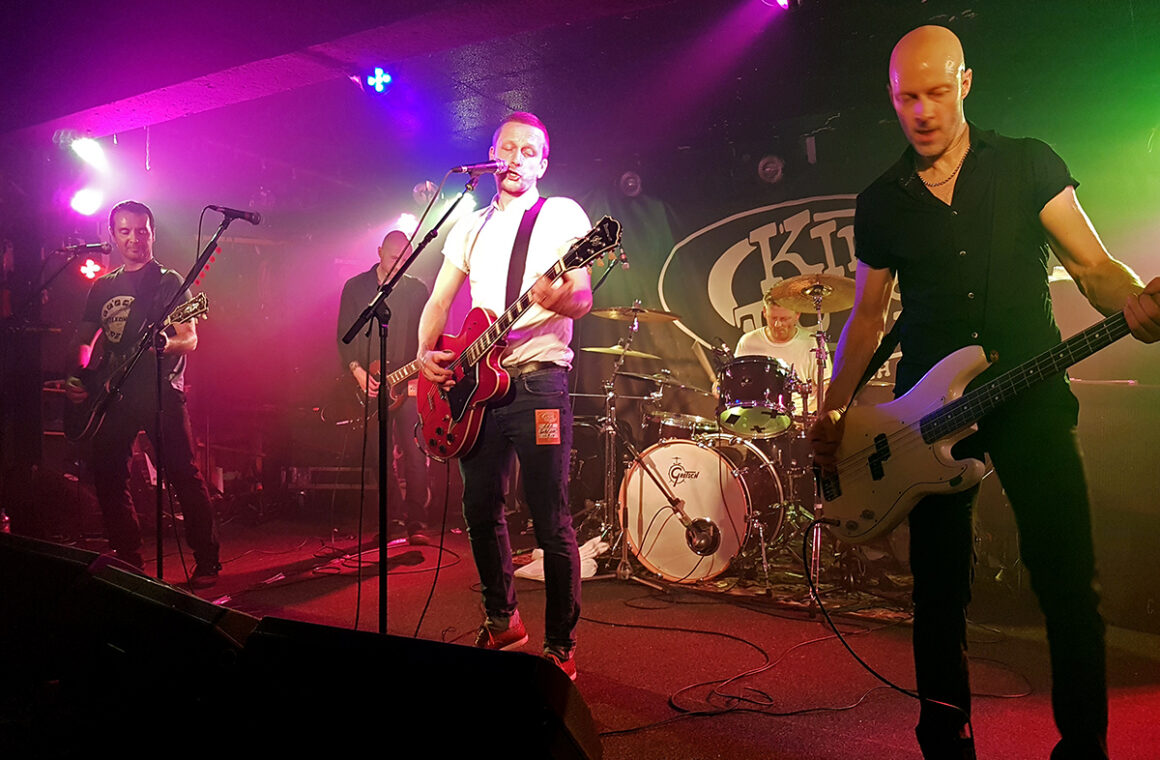 A four-piece band playing under pink and green lights on the low King Tut's stage, with the venue's oval logo on the backdrop