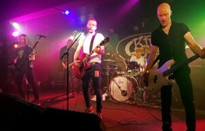 A four-piece band playing under pink and green lights on the low King Tut's stage, with the venue's oval logo on the backdrop