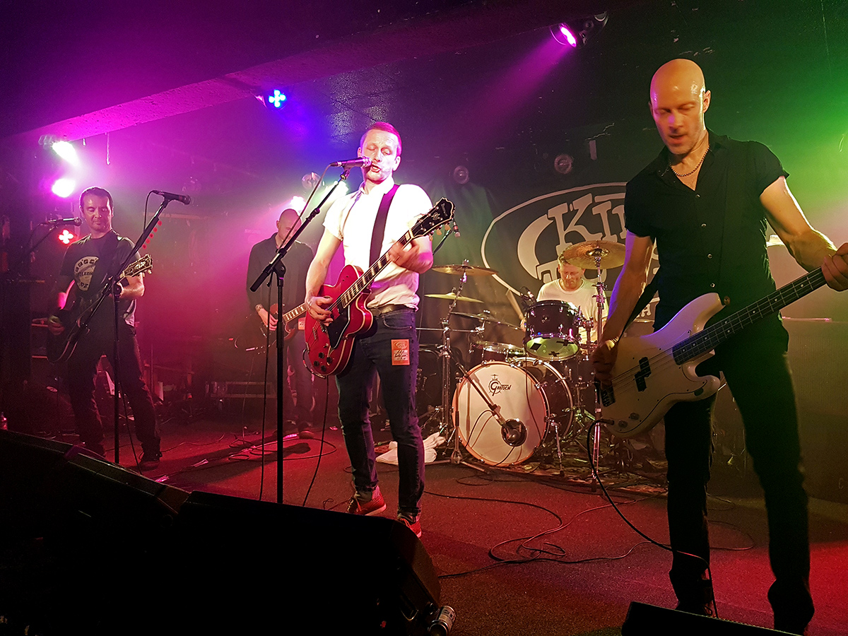 A four-piece band playing under pink and green lights on the low stage at King Tut's Wah Wah Hut