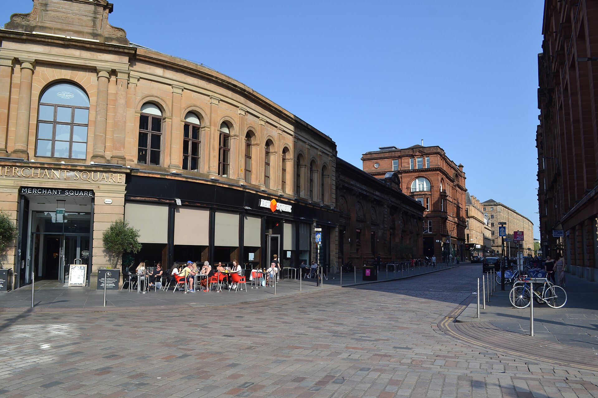 The sandstone Merchant Square building in the Merchant City with its name carved above the entrance, people at red outdoor tables and a cobbled street leading away in spring sunshine