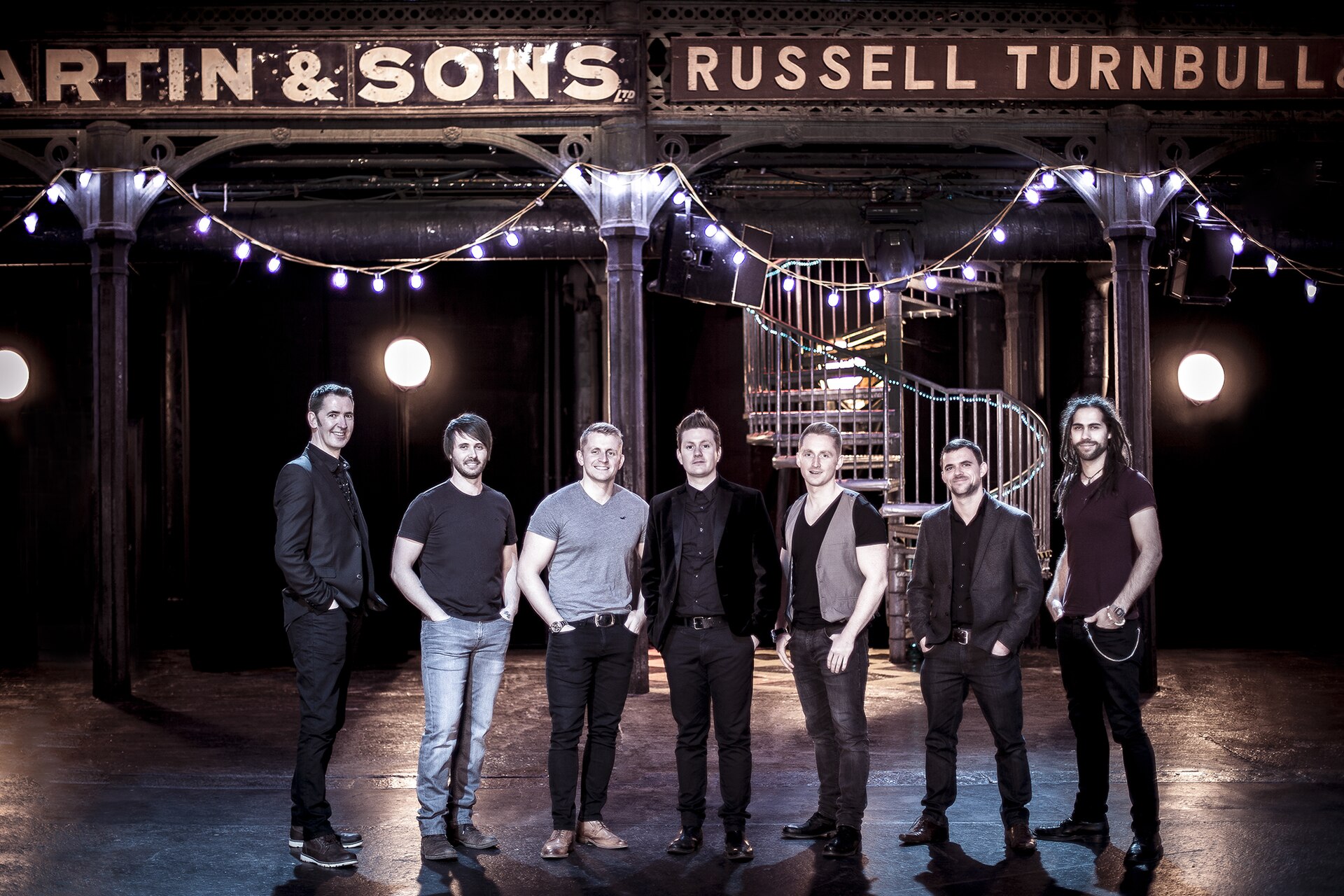 A seven-piece band posing under string lights inside a hall with cast-iron columns, a spiral staircase and old painted fruit merchant signs reading Martin and Sons and Russell Turnbull
