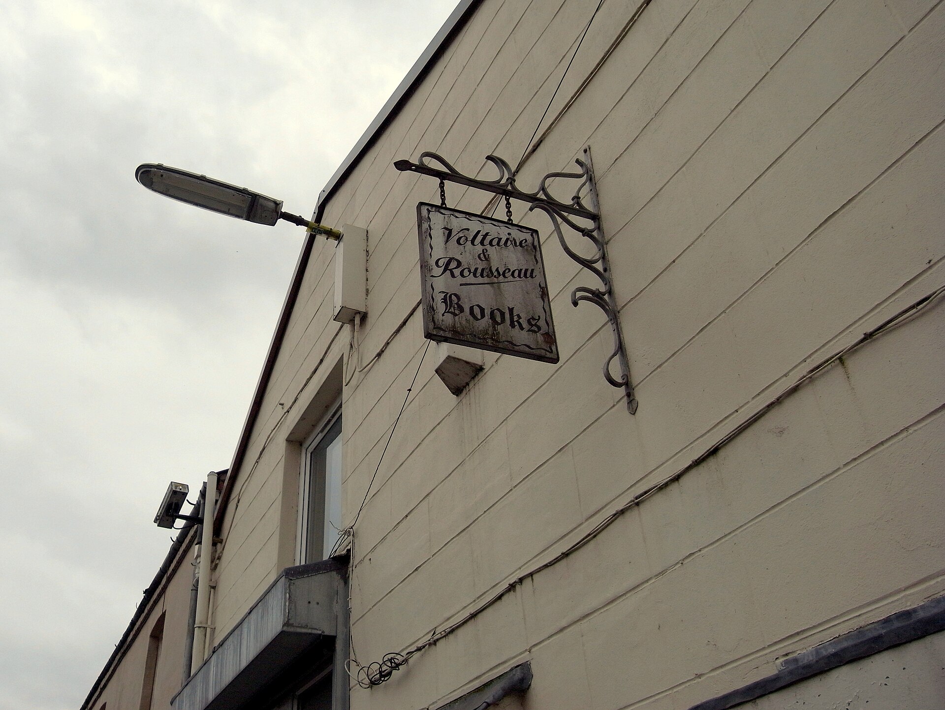 A painted hanging sign for the Voltaire and Rousseau bookshop on a pale building in Otago Lane, under a grey sky