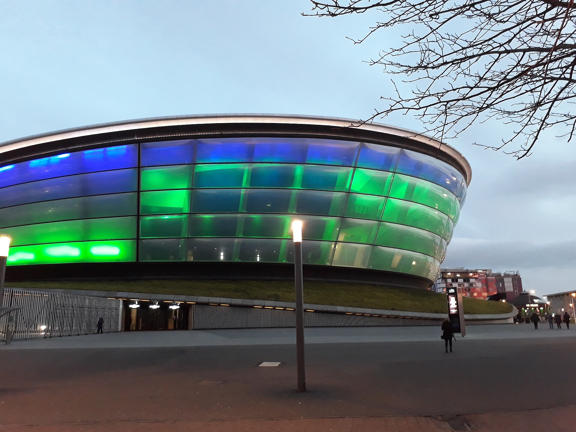 The OVO Hydro lit in blue and green at twilight, seen from the pavement beside the SEC with a lamp post in the foreground