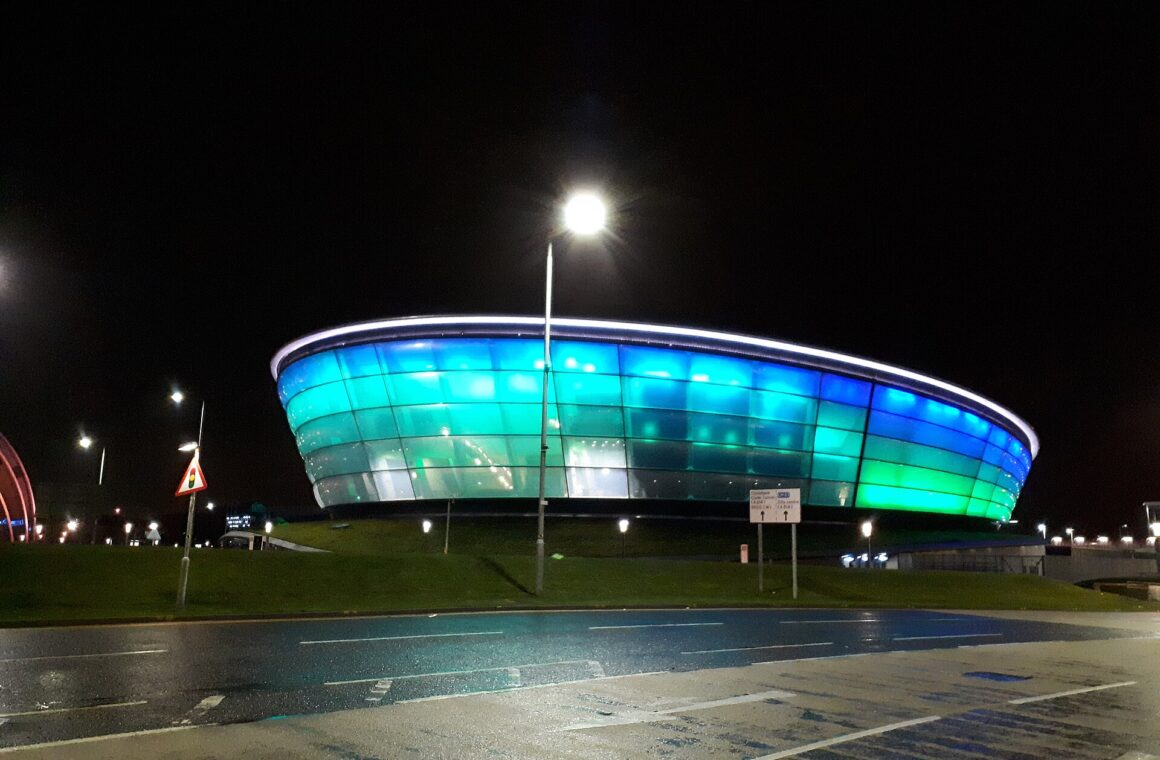 The OVO Hydro arena glowing blue and turquoise at night beside a wet road and street lamps