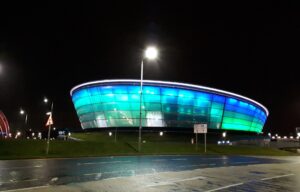 The OVO Hydro arena glowing blue and turquoise at night beside a wet road and street lamps