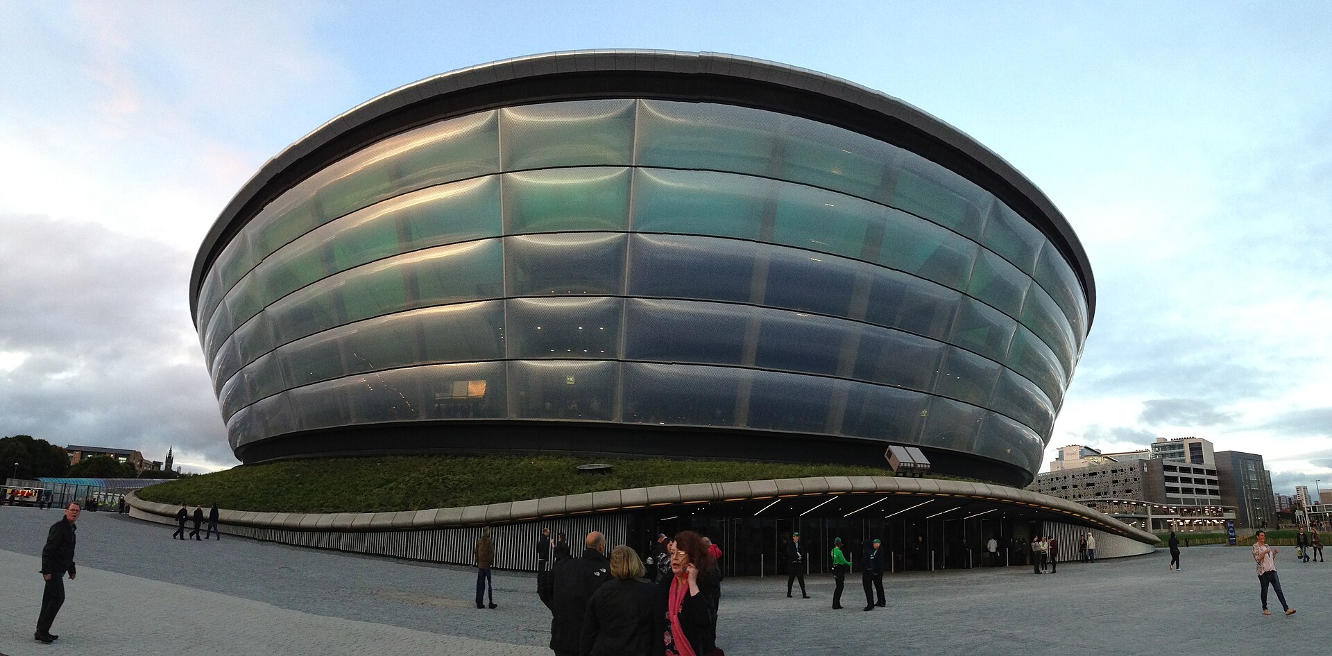 The silver ribbed drum of the OVO Hydro on its grassy plinth at dusk, with people crossing the paved plaza in front
