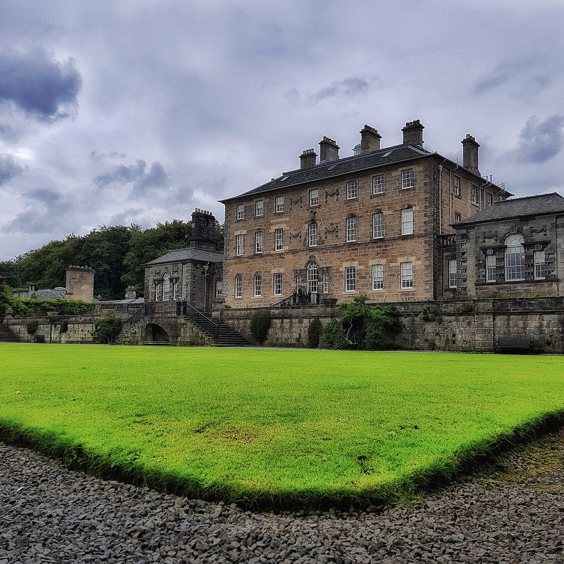 The Georgian stone front of Pollok House above a bright green lawn and terraced garden walls, under a cloudy sky