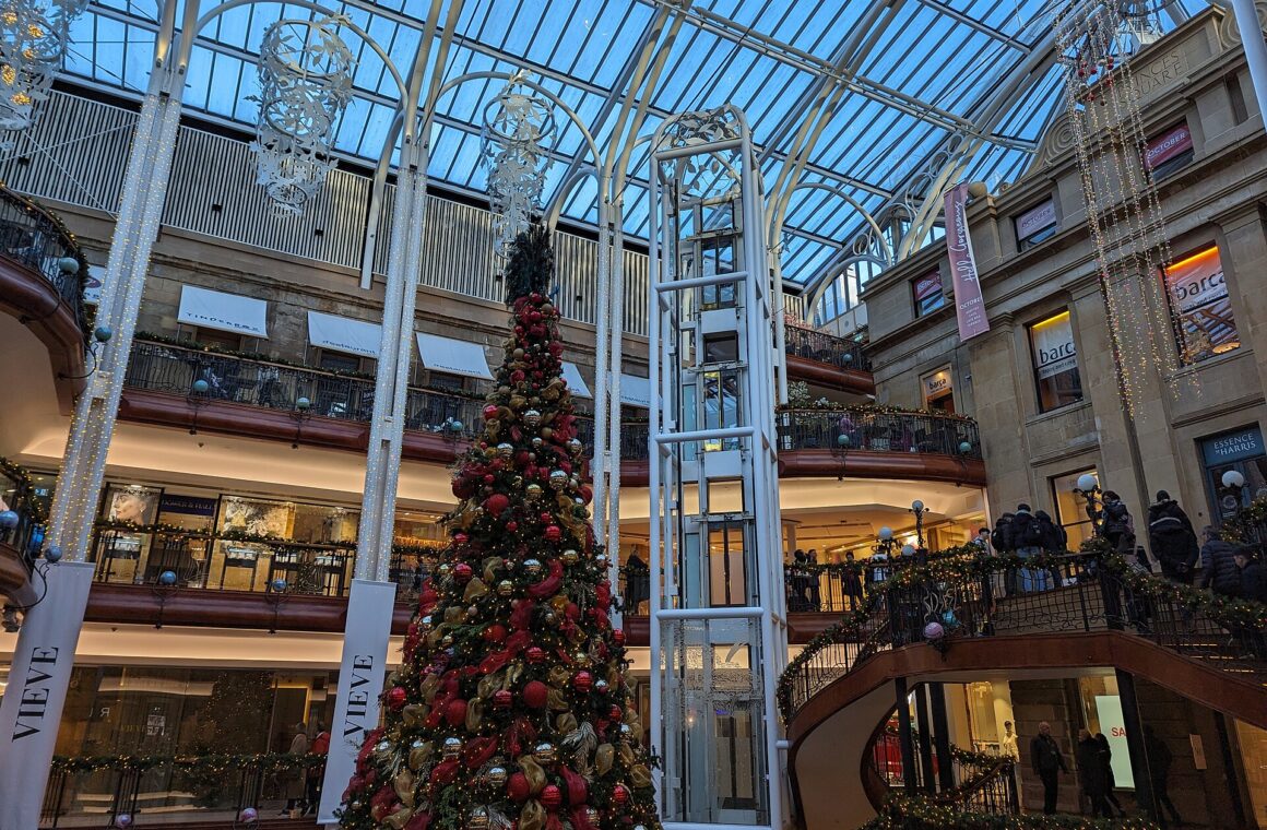 A tall red and gold Christmas tree in the glass-roofed atrium of Princes Square, surrounded by balconies, fairy lights and a curving staircase
