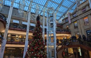 A tall red and gold Christmas tree in the glass-roofed atrium of Princes Square, surrounded by balconies, fairy lights and a curving staircase