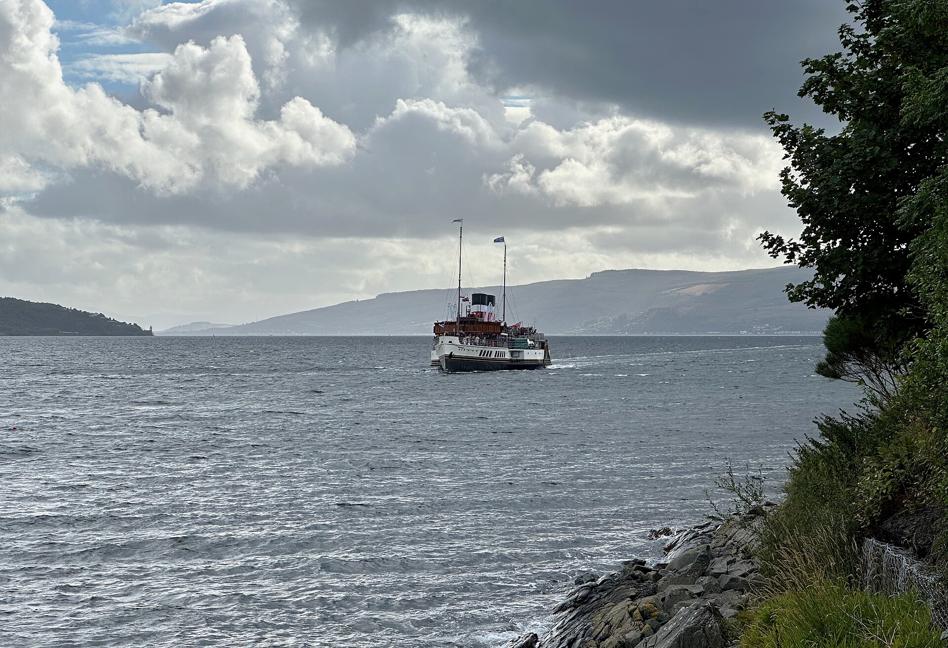 The paddle steamer Waverley crossing the grey-blue Firth of Clyde towards the camera under a sky of big white clouds, with hills on the far shore