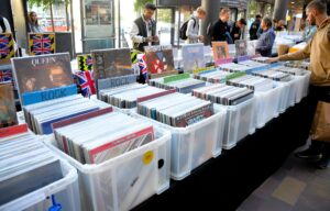 Rows of plastic crates full of LPs at an outdoor record fair, with shoppers browsing along the tables