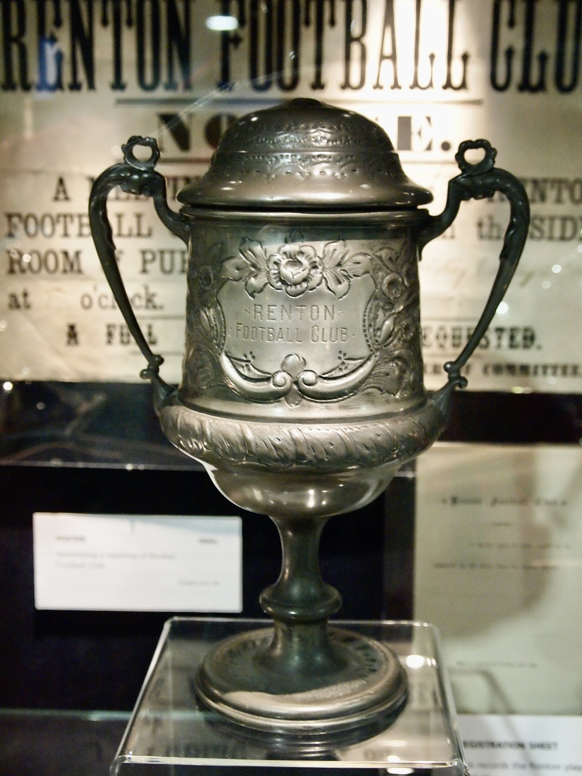 An engraved Victorian silver trophy in a museum display case, inscribed Renton Football Club, with archive documents behind it