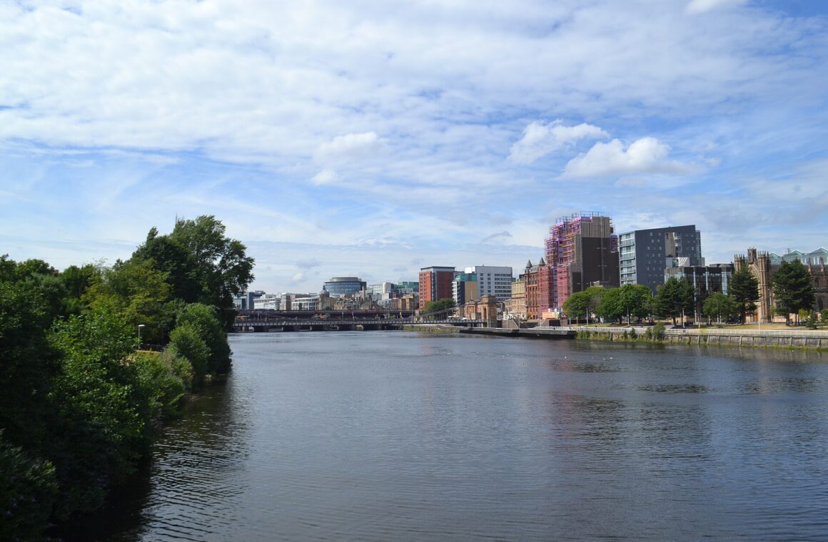 The River Clyde in central Glasgow on a bright July day, with trees in full leaf on one bank, city buildings along the other and railway bridges downstream under a blue sky