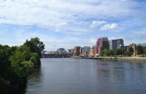 The River Clyde in central Glasgow on a bright July day, with trees in full leaf on one bank, city buildings along the other and railway bridges downstream under a blue sky